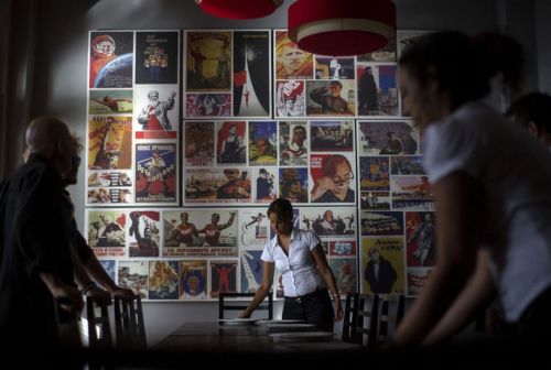 Seorang pelayan restoran menyajikan pesanan makanan dengan latar belakang berbagai propaganda reproduksi poster propaganda Soviet menggantung di dinding di restoran Nazdarovie baru sebagai pelayan mempersiapkan meja untuk latihan pra-peluncuran di Havana, Kuba.